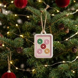 May include: A rectangular Christmas tree ornament with a white border and a beige center, embroidered with colorful circles. The ornament hangs from a natural twine string against a backdrop of a decorated Christmas tree with lights and ornaments.