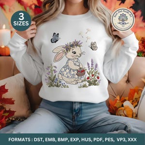 Puede incluir: Sudadera blanca con un conejo bordado con una corona y un vestido floral, sosteniendo una cesta de bayas. Mariposas y flores rodean al conejo. El texto "3 SIZES" es visible. De Knot And Needle Art Embroidery Workshop.