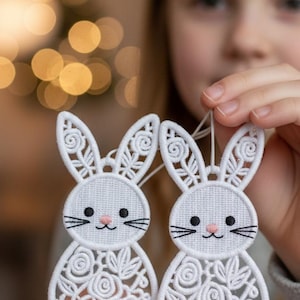 以下が含まれることがあります： 白い刺繍のウサギのオーナメント2つ。花柄と渦巻き模様があります。各ウサギは黒いひげ、目、ピンクの鼻を持っています。オーナメントは紐で吊り下げられており、すぐに飾ることができます。