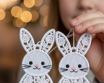 May include: Two white embroidered bunny ornaments with floral and swirl patterns. Each bunny has black whiskers, eyes, and a pink nose. The ornaments are held up by a string, ready to hang.