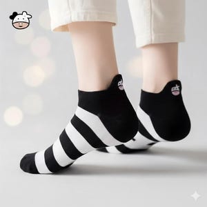 May include: Black and white striped ankle socks. The socks have a black heel and toe, with a white and black striped pattern on the foot. A cartoon cow face is on the back of the sock.