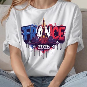 K&ouml;nnte beinhalten: Wei&szlig;es T-Shirt mit einem Grafikdesign mit dem Wort "FRANCE" in Blau und Rot, mit dem Eiffelturm und dem Jahr "2026". Das Design enth&auml;lt Farbspritzer und dekorative Elemente, die einen lebendigen und auff&auml;lligen Look erzeugen.