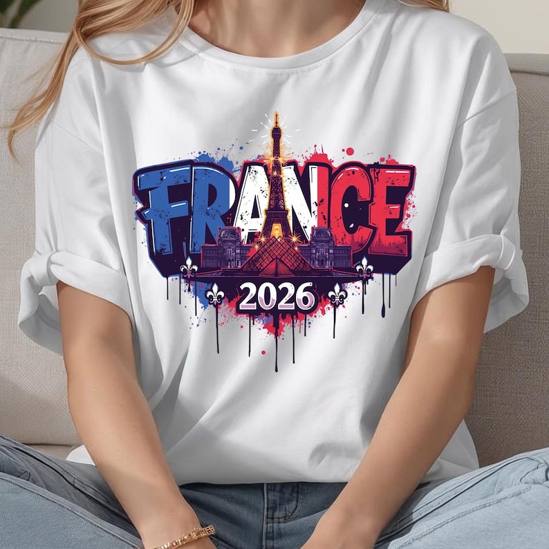 K&ouml;nnte beinhalten: Wei&szlig;es T-Shirt mit einem Grafikdesign mit dem Wort "FRANCE" in Blau und Rot, mit dem Eiffelturm und dem Jahr "2026". Das Design enth&auml;lt Farbspritzer und dekorative Elemente, die einen lebendigen und auff&auml;lligen Look erzeugen.