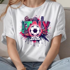 K&ouml;nnte beinhalten: Wei&szlig;es T-Shirt mit einem farbenfrohen Grafikdesign. Das Design zeigt das Wort "ITALY", einen Fu&szlig;ball und das Jahr "2026". Das Design enth&auml;lt italienische Wahrzeichen und Farbspritzer in Pink, Gr&uuml;n und Rot.