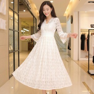 Puede incluir: Un vestido de encaje blanco con escote en V y mangas largas acampanadas. El vestido tiene una cintura ajustada y una falda fluida. El vestido est&aacute; hecho de una delicada tela de encaje y es adecuado para ocasiones especiales.