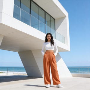 May include: A woman wearing a white long-sleeve crop top and high-waisted, wide-leg rust-colored pants. She is wearing white sneakers. The background features a modern white building with large windows and a view of the ocean under a clear blue sky.