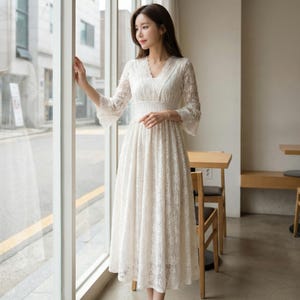 Puede incluir: Un vestido blanco de encaje con escote en V y mangas largas. El vestido tiene un corpi&ntilde;o ajustado y una falda fluida. Las mangas tienen un dise&ntilde;o acampanado. El vestido es hasta la rodilla y parece estar hecho de una delicada tela de encaje.