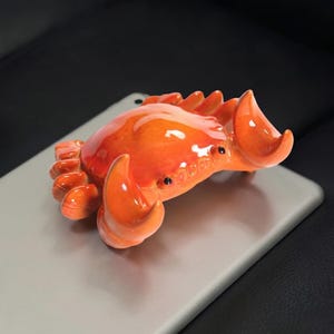 May include: A glossy, orange crab figurine with black eyes and detailed claws. The crab is resting on a light gray surface, showcasing its vibrant color and intricate design. This decorative item adds a touch of coastal charm.