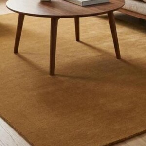 May include: A circular, dark brown wooden coffee table with three legs, a book, and a vase. The table is on a large, rectangular, solid brown rug. The rug is made of a soft material.