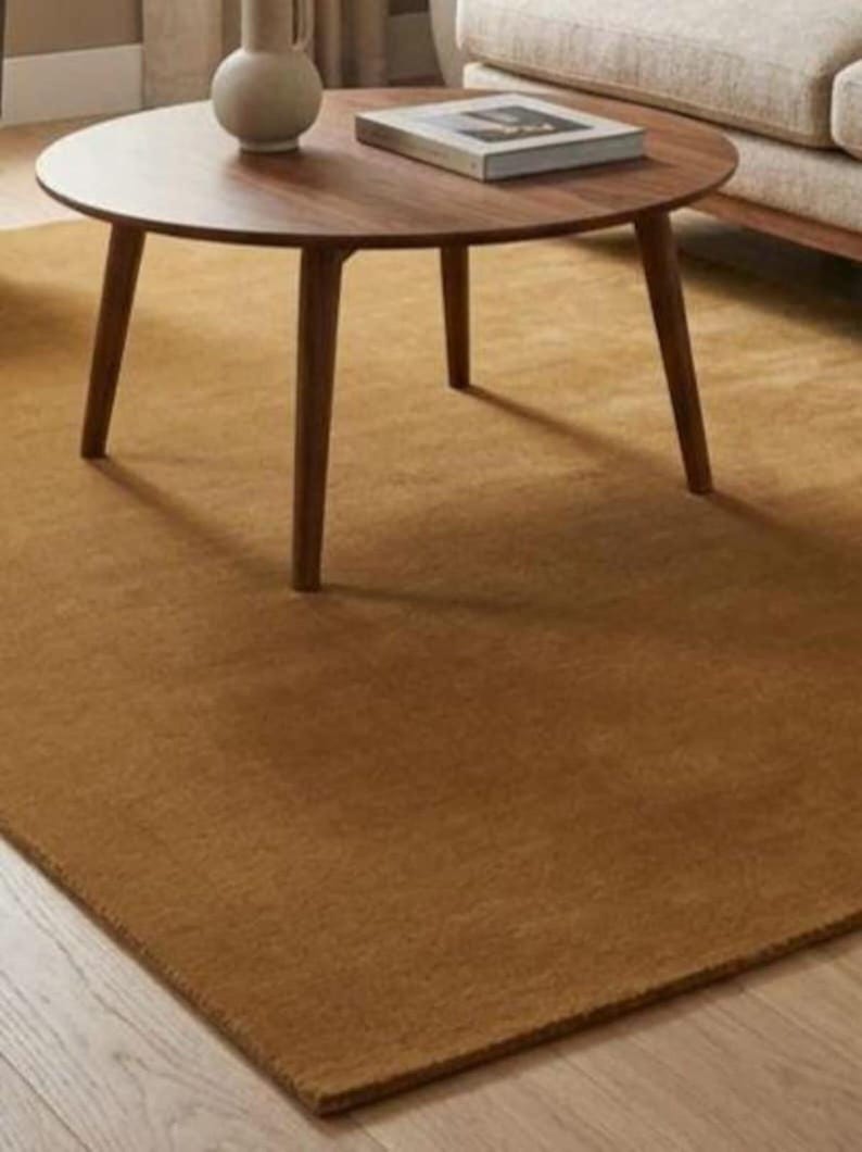 May include: A circular, dark brown wooden coffee table with three legs, a book, and a vase. The table is on a large, rectangular, solid brown rug. The rug is made of a soft material.