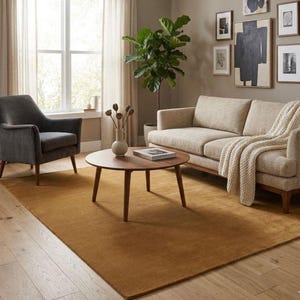 May include: A living room with a mustard-coloured rug, a light-coloured sofa with a cream throw, and a dark grey armchair. A round wooden coffee table is in the centre, and framed artwork decorates the wall. A large green plant adds a natural element.