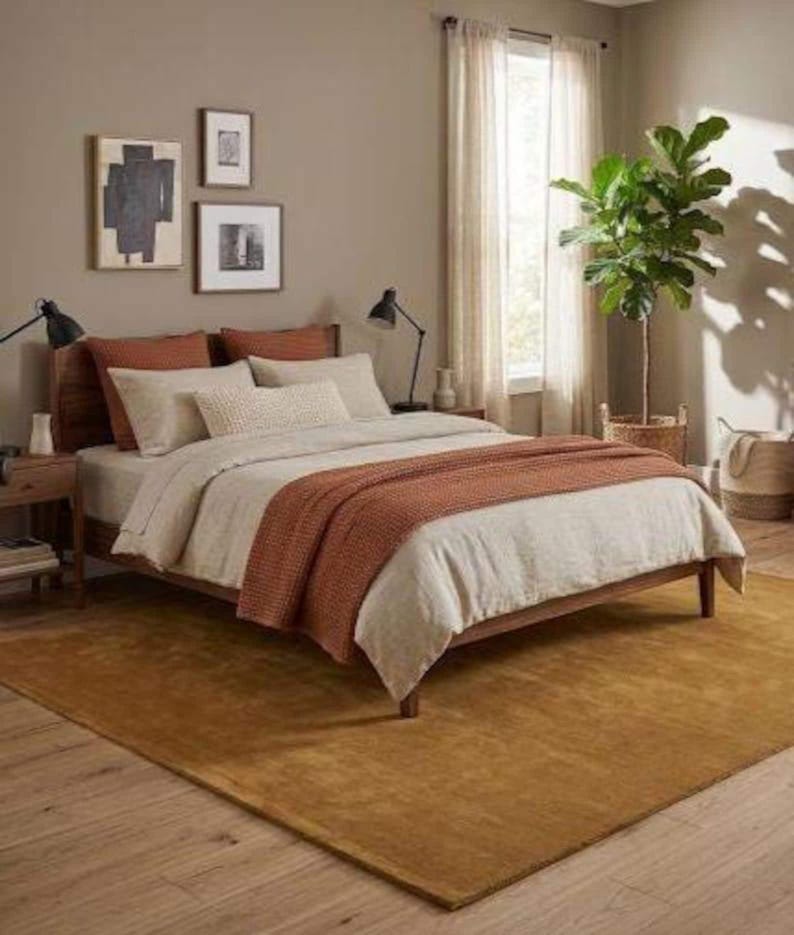 May include: A bedroom with a wooden bed, white bedding, and a rust-coloured throw. The room has a mustard-coloured rug, framed art, and a potted plant. Two black lamps are on the bedside tables. The rug covers an area of approximately 2.5 x 3.5 meters.