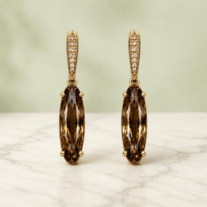 May include: A pair of gold-coloured drop earrings. Each earring has a brown, oval gemstone hanging from a gold hook decorated with small, clear stones. The earrings are displayed on a white marble surface against a soft green background.