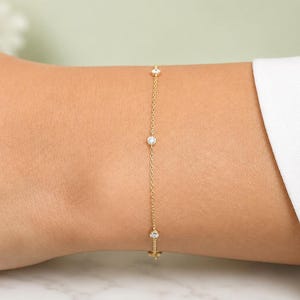 May include: A delicate gold bracelet featuring three clear, round gemstones. The bracelet is attached to a thin gold chain and is worn on a wrist. The bracelet is a piece of jewelry.