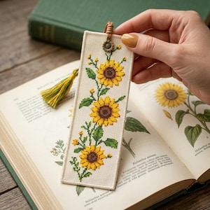 May include: A rectangular bookmark made of cream-colored fabric featuring a cross-stitched pattern of three yellow sunflowers with green leaves and stems. The top includes a small brown leather loop with a metal sunflower charm and a yellow tassel.
