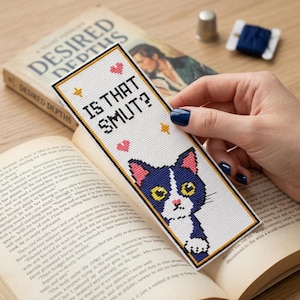 May include: A rectangular cross-stitch bookmark featuring a white background with a black, white, and pink cat illustration at the bottom. The text "IS THAT SMUT?" is stitched in black letters, accented by small pink hearts and yellow stars.