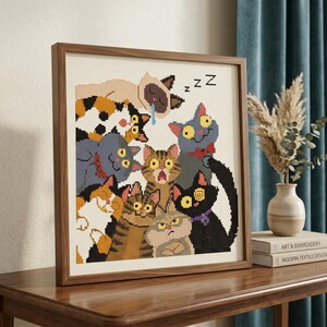 May include: A framed cross-stitch embroidery featuring a group of nine cartoon cats with various expressions and colours, including a sleeping Siamese cat with a blue drool drop and the letter Z floating above, all set against a cream background.