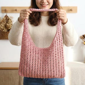 May include: A light pink, open-weave crochet tote bag with a shoulder strap. The bag is held up by a person. The bag's design features a series of evenly spaced holes, creating a textured and airy appearance. The bag is a soft pink color.