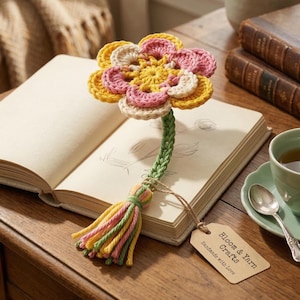 May include: A handmade crochet flower bookmark with a yellow, pink, and cream flower head, green stem, and a tassel. The bookmark is placed in an open book on a wooden table. A tag reads "Bloom & Yarn Crafts, Handmade with Love."
