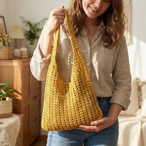 May include: A mustard yellow crochet tote bag with long straps. The bag is made of a loosely woven material, creating a net-like appearance. The bag is held by a person wearing a light-colored button-down shirt and jeans.