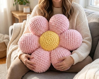 Op de afbeelding: Een bloemvormig kussen met een felgeel rond midden omringd door zes afgeronde, zachtroze bloemblaadjes. Het kussen is gemaakt van dik, grof gebreid garen met een zichtbaar, getextureerd lussenpatroon over het gehele oppervlak.