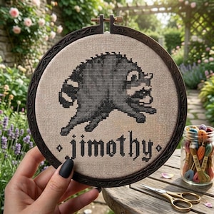 May include: A round wooden embroidery hoop containing a beige fabric panel with a black cross-stitched raccoon graphic and the text "jimothy" in a gothic font. The hoop features a dark brown frame with an ornate, carved decorative pattern.