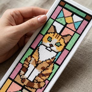 以下が含まれることがあります： オレンジと白の猫のクロスステッチデザインが施された長方形のしおり。猫はピンク、緑、黄色、青の色合いの幾何学的なステンドグラス模様で縁取られており、白い布の縁に黒い糸で輪郭が描かれています。