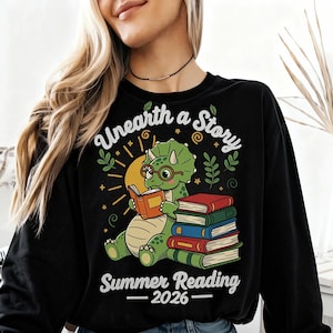 May include: Black sweatshirt with a cartoon dinosaur reading a book, wearing glasses, and sitting in front of a stack of books. The text "Unearth a Story" and "Summer Reading 2026" are printed on the shirt.