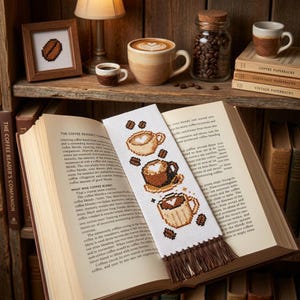 May include: A white fabric bookmark featuring a cross-stitch pattern of three coffee cups with heart-shaped foam designs and scattered coffee beans, finished with a brown fringe at the bottom, resting inside an open hardcover book.