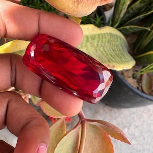 May include: A faceted, rectangular, deep red gemstone. The gem is held between fingers, with a blurred background of green and yellow foliage. The gem's cut creates multiple reflective surfaces, enhancing its brilliance.
