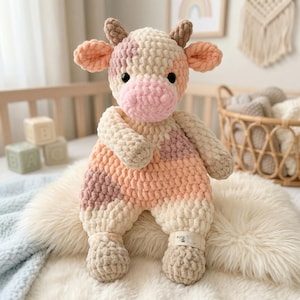 May include: A handmade crochet cow plush toy in soft pastel colors. The cow features a pink nose, ears, and patches, with a cream and peach body. It has small brown horns and black eyes. The plush is sitting on a white faux fur blanket.