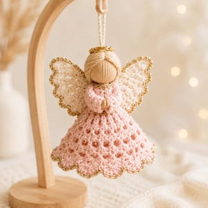 Puede incluir: Un adorno de ángel tejido a ganchillo con un vestido rosa claro de dobladillo festoneado, alas color crema con ribete dorado y una aureola dorada. La figura tiene una cabeza envuelta en hilo y manos juntas en oración, colgada de un cordel.