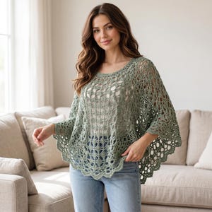 May include: A sage green crochet poncho featuring an intricate openwork pattern with a scalloped hemline. The garment has a wide, rounded neckline and a loose, flowing silhouette that drapes over the shoulders and arms, worn over light blue jeans.