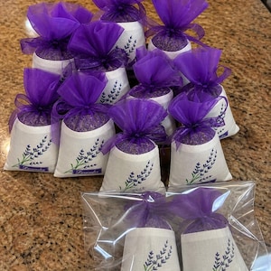 Könnte beinhalten: Eine Sammlung von Lavendelsäckchen. Jedes Säckchen ist ein kleiner, weißer Beutel mit einem Lavendelzweig-Design und dem Wort "Lavender" aufgedruckt. Die Beutel sind mit lila Tüll bedeckt und mit einem Band gebunden. Einige sind in einer transparenten Plastiktüte verpackt.