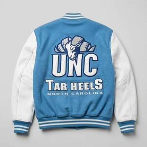 Puede incluir: Una chaqueta universitaria azul claro con mangas blancas y ribetes a rayas. La parte posterior presenta un gran gr&aacute;fico bordado de una cabeza de carnero sobre el texto UNC, Tar Heels y North Carolina en letras blancas y azul marino.