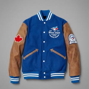 Op de afbeelding: Een blauwe varsity-jas met bruine mouwen, voorzien van een wit gestreepte kraag, boorden en zoom. Op de borst staat het logo van de Blue Jays Baseball Club met de tekst Est. 1977. Een rode esdoornbladpatch bevindt zich op de rechtermouw.