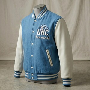 Puede incluir: Una chaqueta universitaria de color azul claro con mangas blancas, que presenta el logotipo y el texto UNC Tar Heels bordados en el pecho. La chaqueta tiene cierre frontal con botones a presi&oacute;n, pu&ntilde;os y cuello de canal&eacute; a rayas y dos bolsillos laterales.