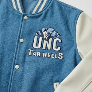 Puede incluir: Una chaqueta universitaria de mezcla de lana azul con mangas de cuero blanco y cuello acanalado a rayas. El pecho presenta un logotipo bordado de UNC Tar Heels con un gr&aacute;fico de carnero en hilo blanco, azul marino y azul claro, con broches.