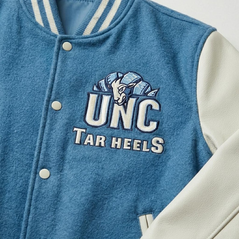 Puede incluir: Una chaqueta universitaria de mezcla de lana azul con mangas de cuero blanco y cuello acanalado a rayas. El pecho presenta un logotipo bordado de UNC Tar Heels con un gr&aacute;fico de carnero en hilo blanco, azul marino y azul claro, con broches.