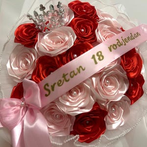 Pode incluir: Um buquê de rosas de cetim vermelhas e rosa claro com um pequeno ornamento de coroa prateada. Uma fita rosa claro está disposta sobre as flores com o texto dourado Sretan 18 rodjendan e um grande laço de cetim rosa na base.