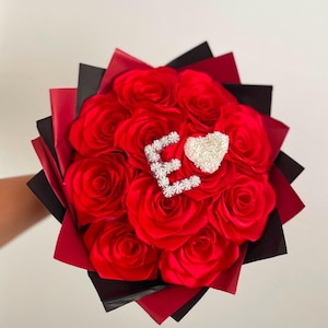 Può includere: Un bouquet di rose artificiali rosso vivo avvolto in strati di carta nera e rossa. Al centro delle rose si trovano una lettera E bianca testurizzata e un ornamento bianco a forma di cuore, entrambi con una finitura scintillante e cristallina.
