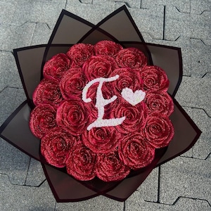 Könnte beinhalten: Ein Strauß aus roten Glitzerrosen, eingewickelt in dunkles, durchscheinendes Papier. In der Mitte der Rosen befinden sich ein großer weißer Buchstabe E und ein kleines weißes Herz. Das Arrangement ist kreisförmig und hat viele Blütenblätter.