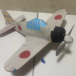 Könnte beinhalten: Ein Modellflugzeug mit grauem und schwarzem Rumpf, blauen Cockpitfenstern und einem Propeller. Die Flügel weisen rote Kreise auf und das Heck trägt die Nummer "AI-105". Das Flugzeug ist eine Nachbildung eines Oldtimer-Flugzeugs.
