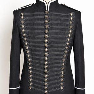 Peut inclure: Veste noire de style militaire avec un col montant et des boutons argentés. La veste présente des détails décoratifs en corde et une bordure blanche sur le col et les poignets. La veste est présentée sur un mannequin.