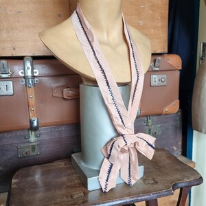 May include: A vintage blush pink necktie with dark blue stitching detail. The tie is draped on a mannequin bust. The tie is tied in a bow at the bottom. The background includes antique luggage and a wooden table.