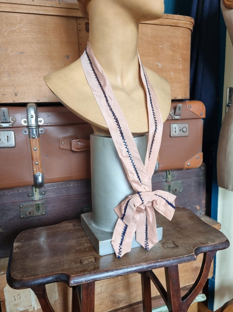 May include: A vintage blush pink necktie with dark blue stitching detail. The tie is draped on a mannequin bust. The tie is tied in a bow at the bottom. The background includes antique luggage and a wooden table.