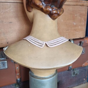 Peut inclure: Un buste de mannequin vintage avec des cheveux bruns et un col rose clair. Le mannequin est expos&eacute; sur un support argent&eacute;, avec un buste de couleur beige. L'arri&egrave;re-plan comprend des bagages anciens.