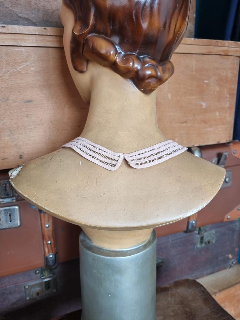 Peut inclure: Un buste de mannequin vintage avec des cheveux bruns et un col rose clair. Le mannequin est expos&eacute; sur un support argent&eacute;, avec un buste de couleur beige. L'arri&egrave;re-plan comprend des bagages anciens.