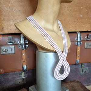 Peut inclure: Un collier rose clair avec un motif tiss&eacute; est pr&eacute;sent&eacute; sur un buste de mannequin. Le collier a un design en boucle et est fait d'un mat&eacute;riau d&eacute;licat. Le buste est mont&eacute; sur un support argent&eacute;, avec une malle marron en arri&egrave;re-plan.