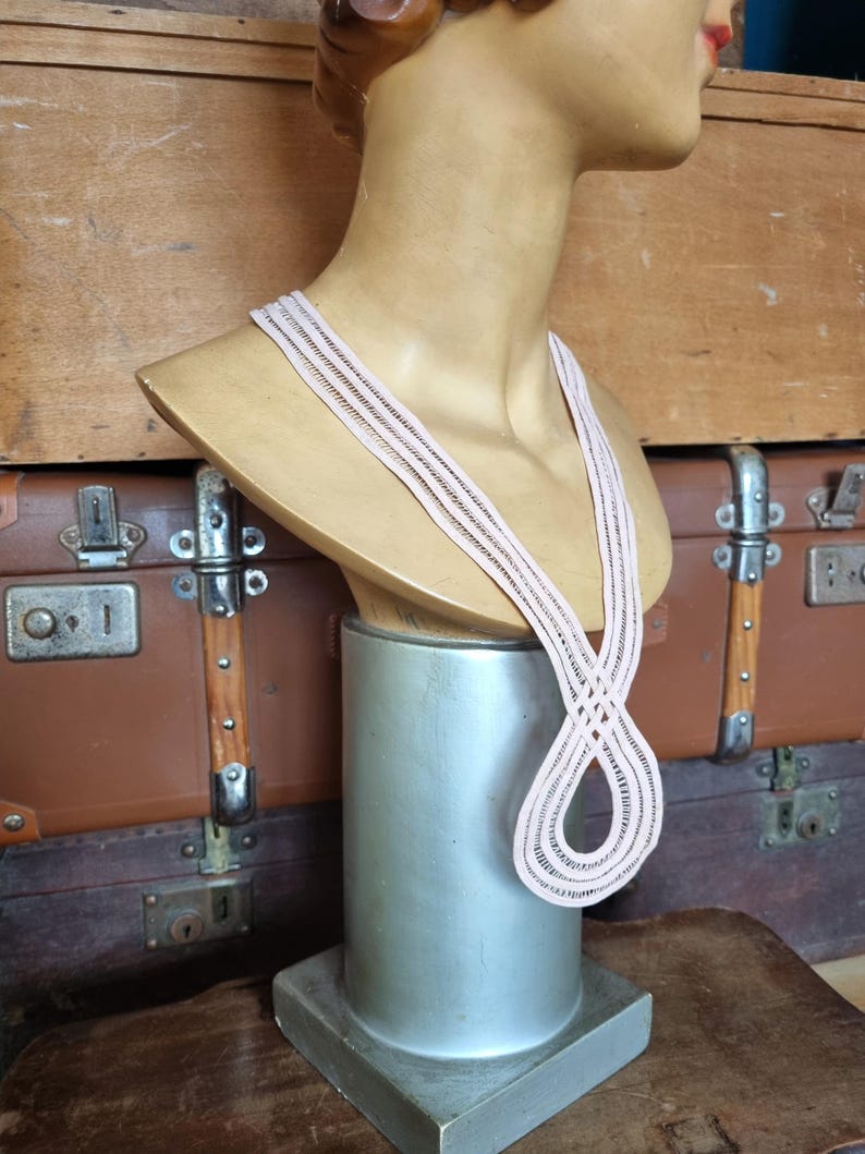 Peut inclure: Un collier rose clair avec un motif tiss&eacute; est pr&eacute;sent&eacute; sur un buste de mannequin. Le collier a un design en boucle et est fait d'un mat&eacute;riau d&eacute;licat. Le buste est mont&eacute; sur un support argent&eacute;, avec une malle marron en arri&egrave;re-plan.